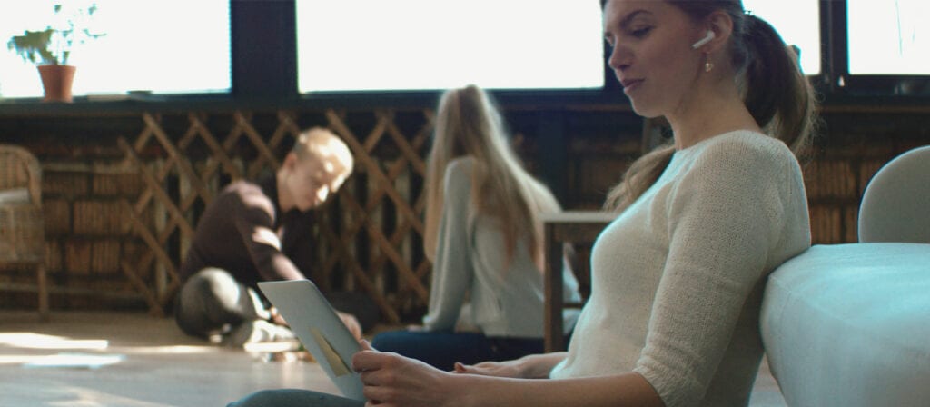 A woman wearing wireless earbuds sits on a sofa, holding a tablet. In the background, two people sit on the floor near a table, working together in a bright room with large windows and bookshelves.