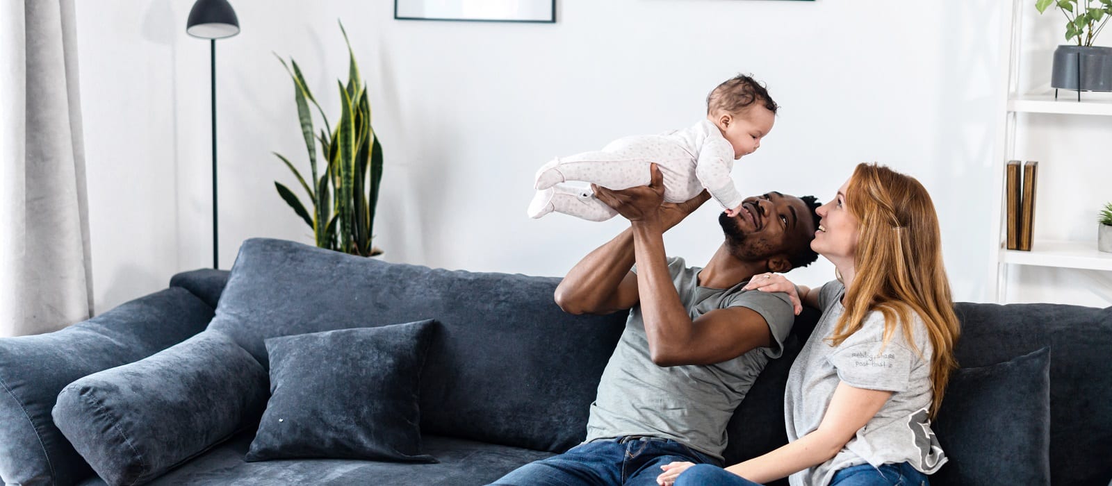 A multiracial family on the comfortable couch at home. A father throwing his baby daughter up, a mother sits near and smiles