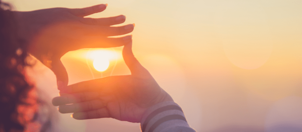 Close-up of two hands forming a rectangular frame with fingers, capturing the sun within the shape at sunset. The background is softly blurred with warm, orange and yellow light.
