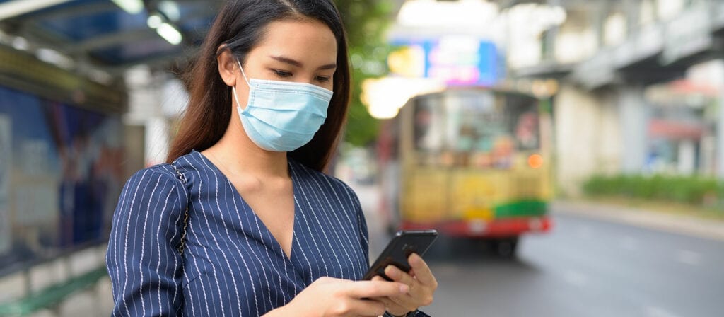 A woman wearing a blue face mask and striped shirt stands at a bus stop, looking down at her smartphone. A bus approaches in the background on a city street.