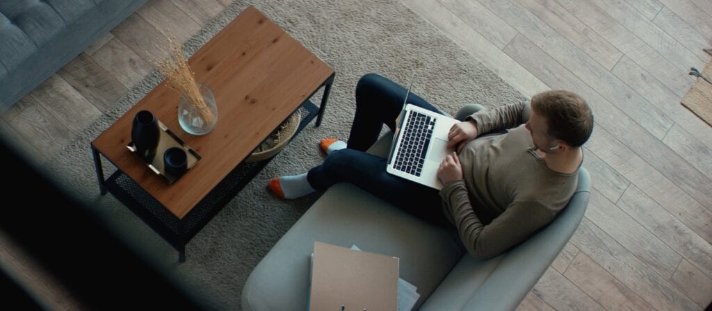 A person sits on a couch using a laptop, with folders and a coffee table nearby. The table holds a vase with dried flowers and two cups. The setting is a cozy, modern living room with wood flooring.