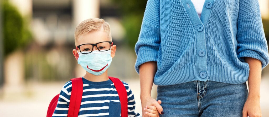 A young child wearing glasses, a striped shirt, a red backpack, and a face mask with a drawn-on smile holds hands with an adult dressed in a blue sweater.
