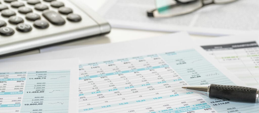 A close-up of financial documents with charts and numbers, a pen resting on one paper, a calculator, and eyeglasses in the background. The scene suggests financial analysis or accounting work.