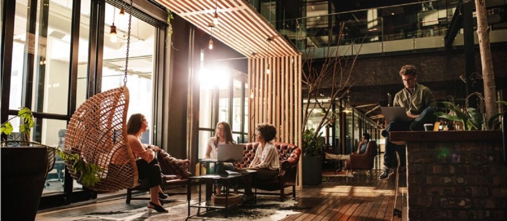 A modern, sunlit office space with four people working and chatting. Three sit on chairs and a hanging seat, one sits on a counter with a laptop. Large windows, plants, and wood accents create a relaxed, collaborative atmosphere.