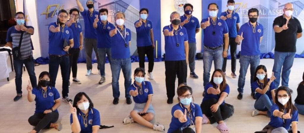 A group of people wearing blue shirts and face masks pose indoors, many standing and some sitting cross-legged on the floor, each holding up their right hand with their index finger extended.