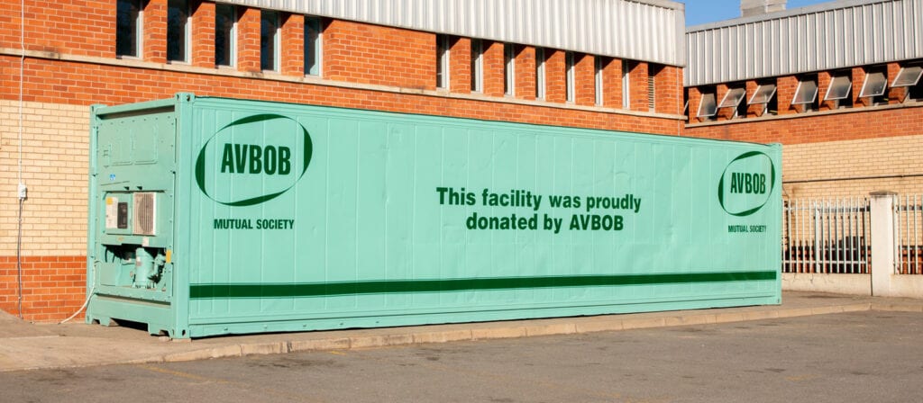 A large mint-green shipping container with the text “This facility was proudly donated by AVBOB” is placed in a parking area outside a brick and cream-colored building.