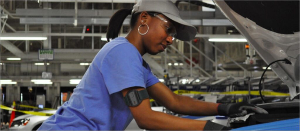 A woman wearing safety glasses, gloves, and a cap works on assembling a vehicle in a factory setting, surrounded by industrial equipment and machinery.