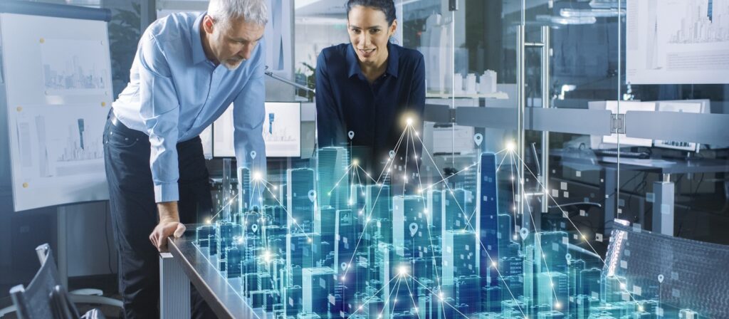 Two professionals examine a digital 3D model of a city displayed on a table, with illuminated holographic buildings and data points, in a modern office setting with architectural plans in the background.
