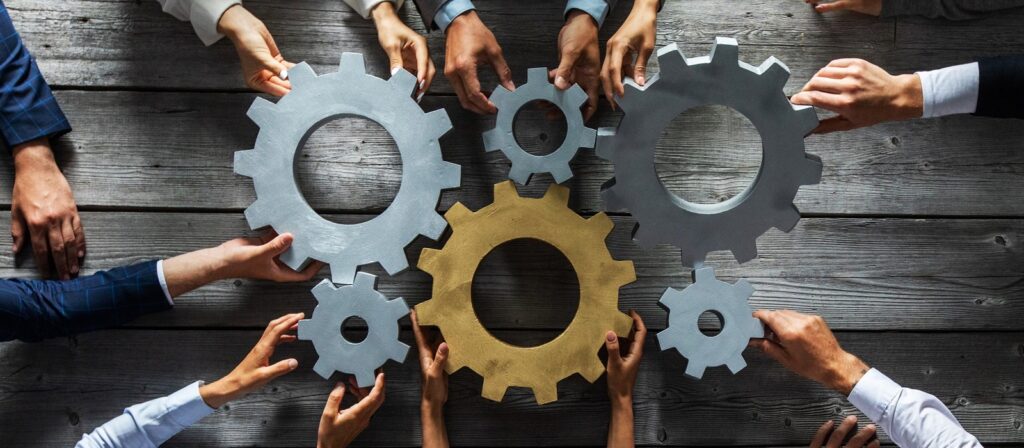 Several hands from different people hold large, interlocking gear wheels above a wooden table, symbolizing teamwork, collaboration, and working together to achieve a common goal.