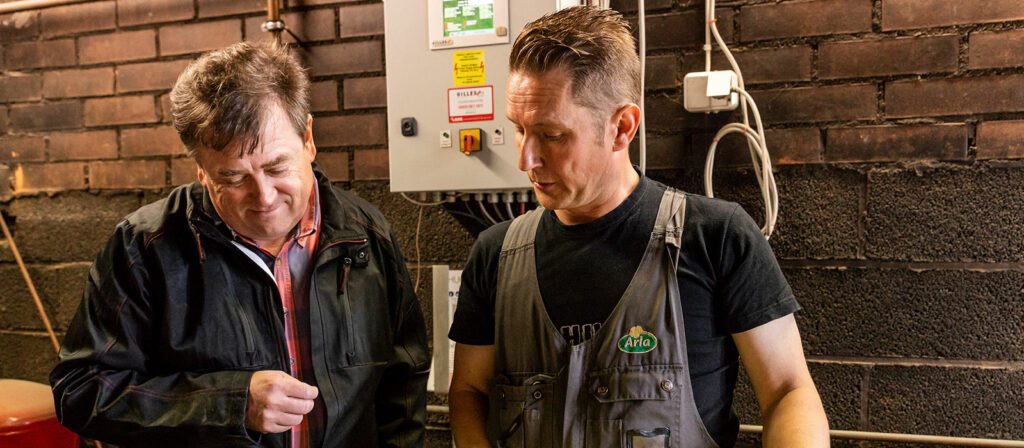 Two men stand indoors near machinery; one wears a black jacket and plaid shirt, the other wears overalls with an Arla logo. They appear to be discussing or inspecting something together. Brick wall and cables are in the background.