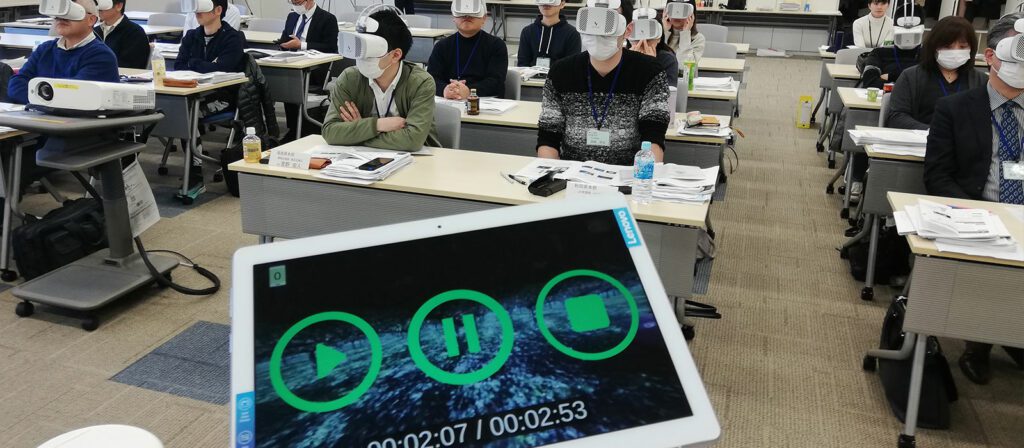 A classroom of people wearing VR headsets sit at desks, facing forward. In the foreground, a tablet displays a timer with play, pause, and stop icons on the screen.