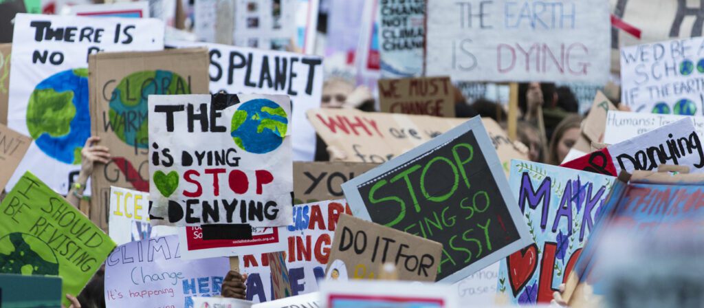 A crowd of people holds up colorful protest signs with messages about climate change, such as The planet is dying, stop denying, The Earth is dying, and Global warming.