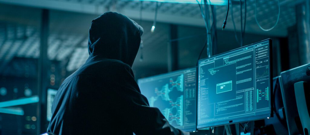 A person wearing a hooded sweatshirt sits in a dark room, facing multiple computer monitors displaying code, maps, and data, suggesting hacking or cybersecurity activity.