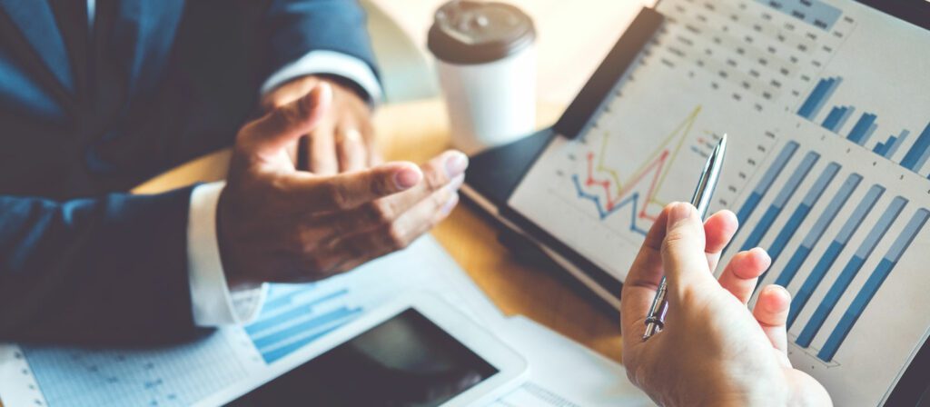 Two people discuss financial charts and graphs displayed on a tablet and printed paper. One person gestures with their hands, while the other holds a pen. A coffee cup and smartphone are on the table.