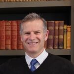 A man with short gray hair, wearing a white shirt, blue tie, and black sweater, smiles in front of a bookshelf filled with old, leather-bound books.