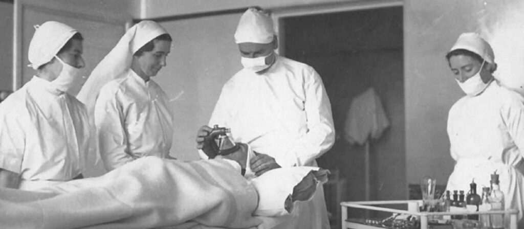 Four medical professionals in early 20th-century attire surround a patient lying on an operating table. The staff wear white gowns, masks, and caps. One doctor appears to administer anesthesia while others observe.