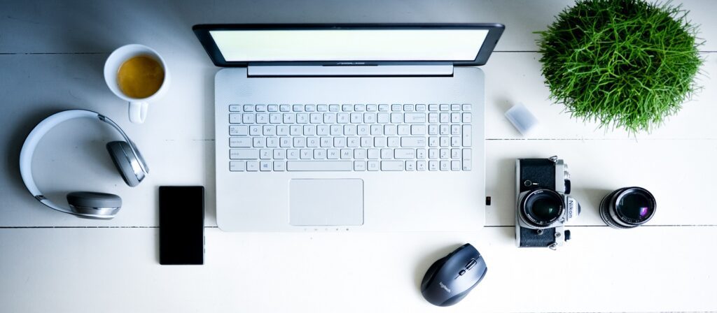 A white laptop on a white desk, surrounded by a cup of coffee, headphones, a smartphone, a computer mouse, a vintage camera with an extra lens, a small plant, and a USB drive.