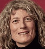 A person with wavy, light-colored hair and a slight smile is shown against a burgundy background. They are wearing a black top.