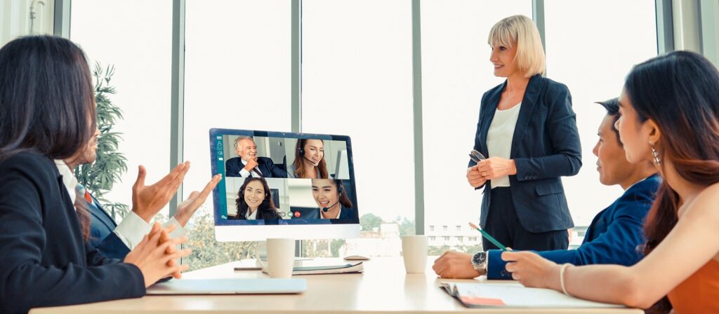 Four people sit around a conference table with laptops, while a woman stands and presents. A computer screen shows four colleagues joining the meeting via video call. Large windows are in the background.