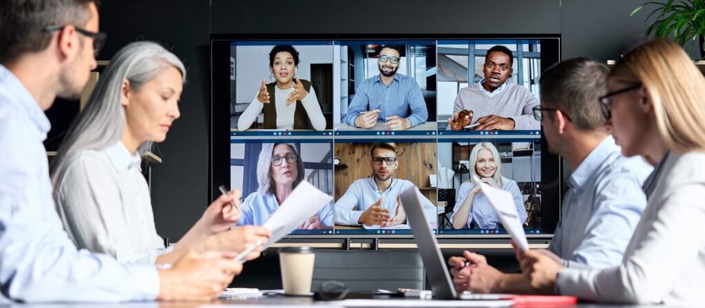 A group of people sit at a table with documents, participating in a video conference with nine colleagues shown on a large screen, all engaged in discussion.
