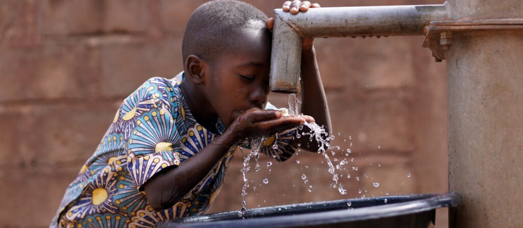 A young boy wearing a colorful patterned shirt drinks water from a hand pump, catching the flowing water in his hands, with a black container below to collect excess water.