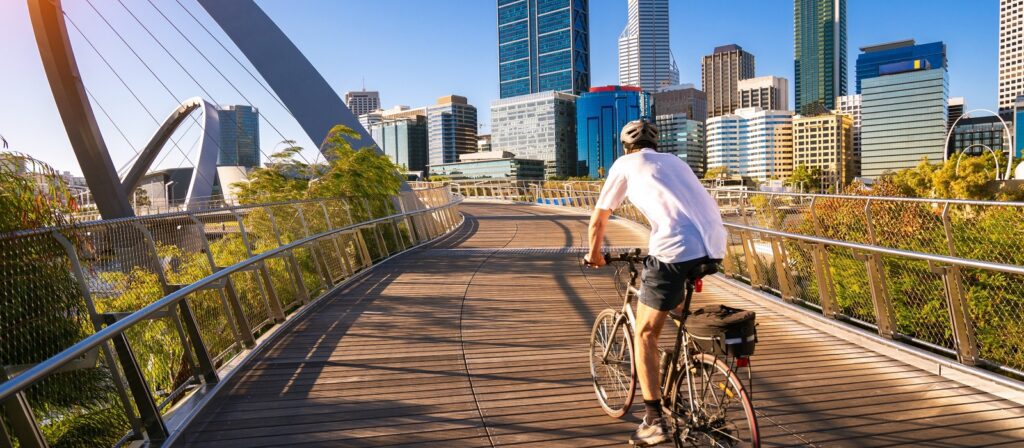 A cyclist wearing a helmet rides on a curved pedestrian bridge toward a modern city skyline on a sunny day, surrounded by greenery and tall buildings.