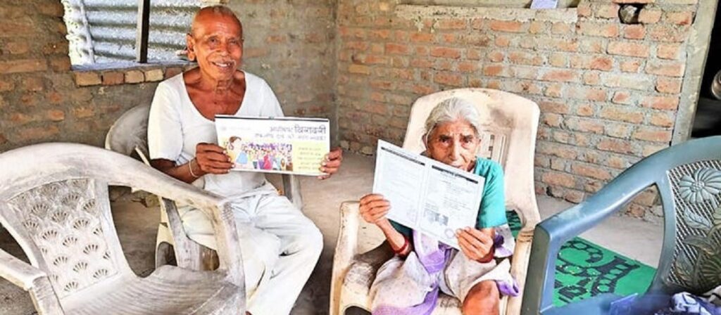 Two elderly people sit on plastic chairs in a rustic room with brick walls, smiling and holding up documents—one a certificate with a photo, the other papers—while empty chairs surround them.