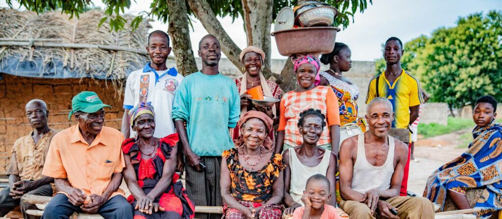 A group of people of various ages, smiling and posing outdoors in front of a tree and rustic buildings. Some are seated, others stand behind them, with colorful clothes and dishes balanced on a woman’s head.