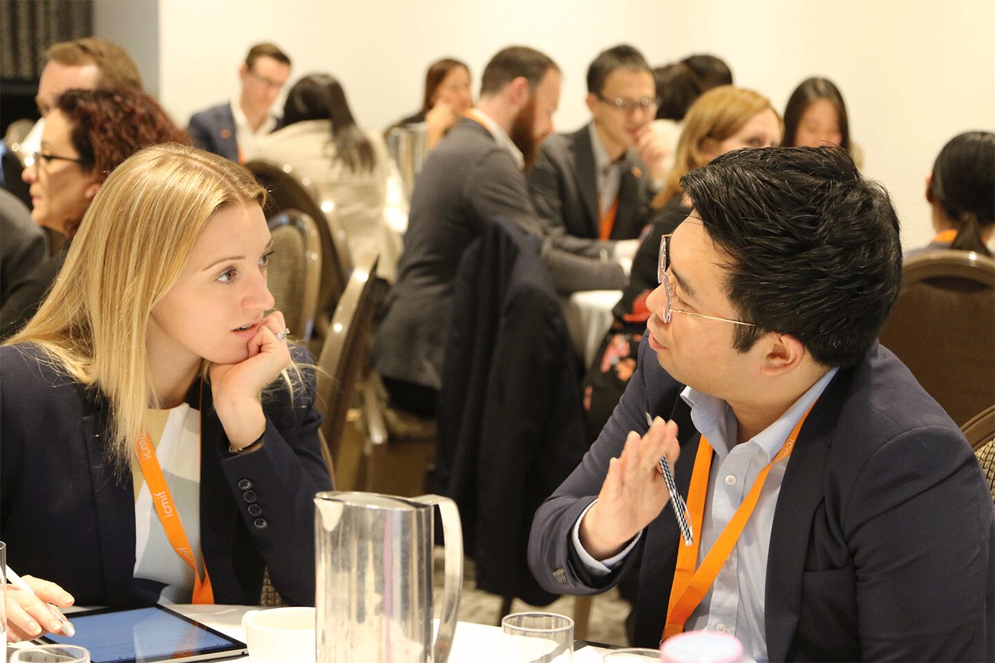 Two people wearing business attire and orange lanyards sit at a table, engaged in a focused conversation at a conference or meeting, with other groups talking in the background.