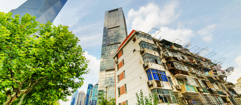 Modern glass skyscrapers tower over an older, worn apartment building with visible balconies and air conditioning units; green tree branches frame the left side, set against a bright, cloudy sky.