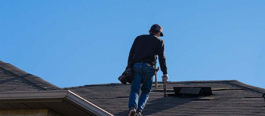 A person wearing a cap, long sleeves, and jeans stands on a house roof with tools, appearing to inspect or repair shingles under a clear blue sky.