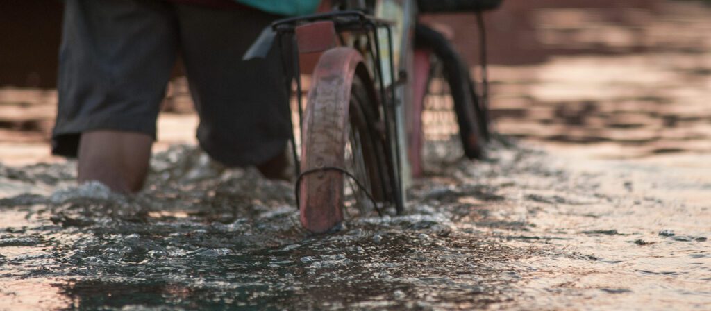 A person wades through knee-deep water, pushing a bicycle. The lower part of their legs and the rear wheel of the bike are visible, with ripples forming around them.
