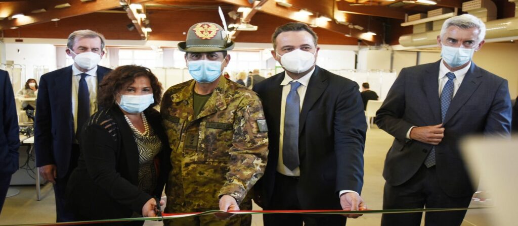 A group of five people, including a military officer in uniform and four individuals in suits, cut a ribbon together at an indoor event. All are wearing face masks.
