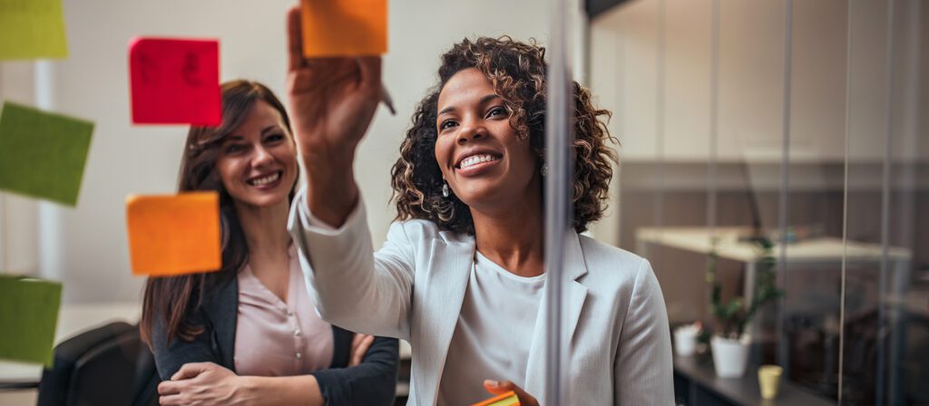 Two women in an office smile as one places a colorful sticky note on a glass wall covered with other notes, suggesting teamwork or a brainstorming session.