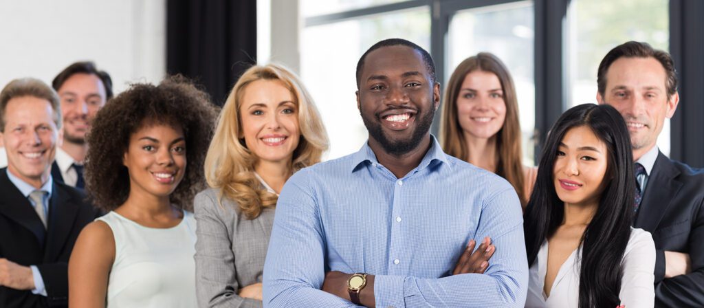 A diverse group of eight professionally dressed people stand together indoors, smiling confidently at the camera. The background shows large windows and a bright, modern office setting.