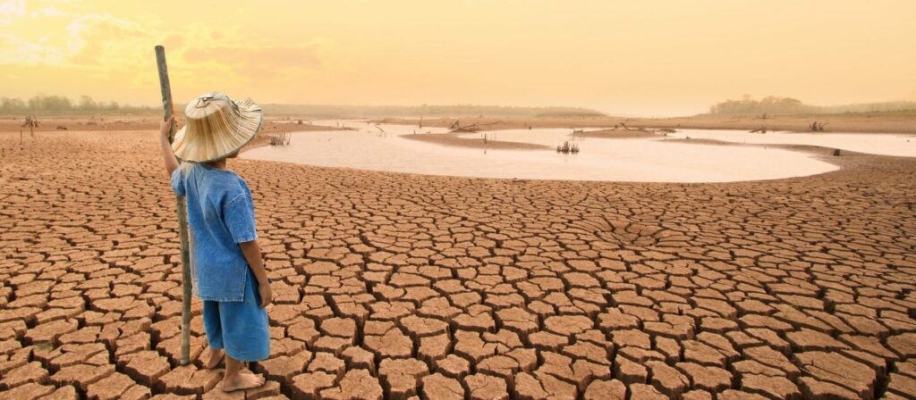 A child wearing a hat and blue clothes stands with a stick on cracked, dry earth near a shrinking water body under an orange sky, illustrating drought and water scarcity.