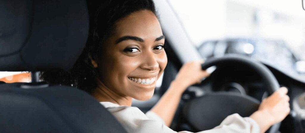 A woman sitting in the driver’s seat of a car, smiling and looking back over her shoulder towards the camera, with her hands on the steering wheel.