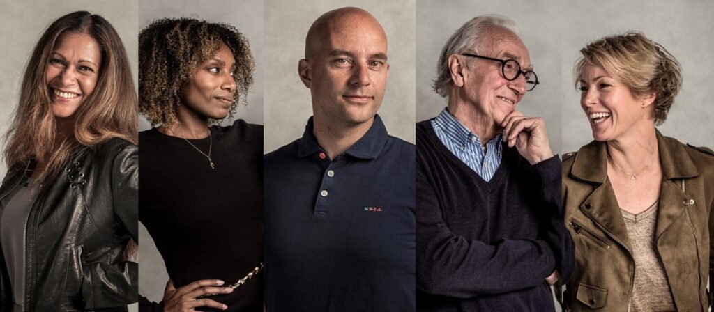 Five adults stand side by side against a neutral background, each facing slightly different directions. They appear happy and engaged, with some smiling and chatting with each other.
