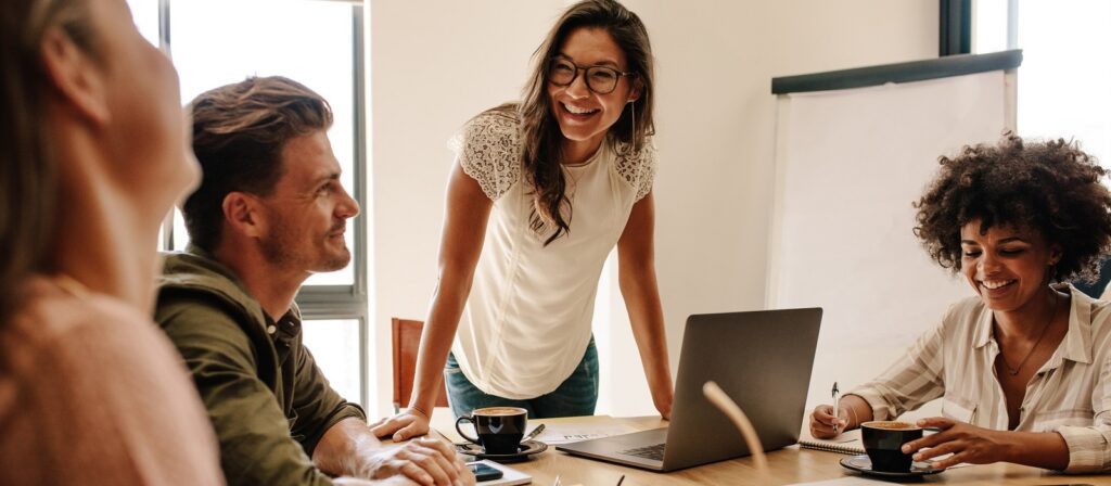 Four people sit around a table in a bright office, smiling and talking. One woman stands, leaning forward with a laptop, while the others sit, drinking coffee and taking notes, suggesting a friendly team meeting.