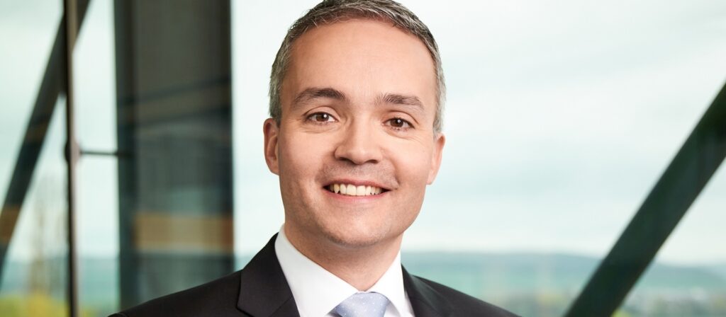 A smiling man in a suit and tie stands indoors with large windows and modern architecture in the background.