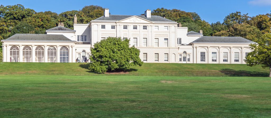 A large, white, neoclassical mansion with tall windows and columns, set on a green lawn with trees and blue sky in the background.