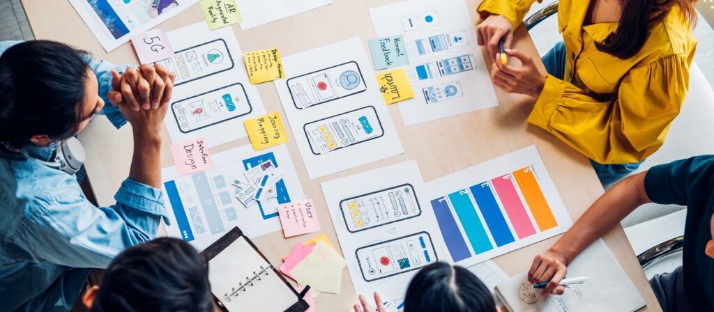Four people sit around a table reviewing mobile app interface sketches, wireframes, and color swatches, with notebooks and post-it notes scattered, suggesting a collaborative design or brainstorming session.