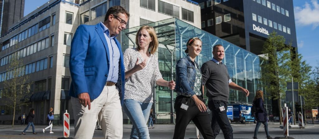 Four people walk together on a city street in front of modern office buildings, one with a visible Deloitte sign. The group appears to be engaged in conversation on a sunny day.