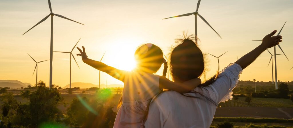 A woman holds a child while both face a sunset, raising their arms toward wind turbines in a green field, symbolizing renewable energy and a hopeful future.