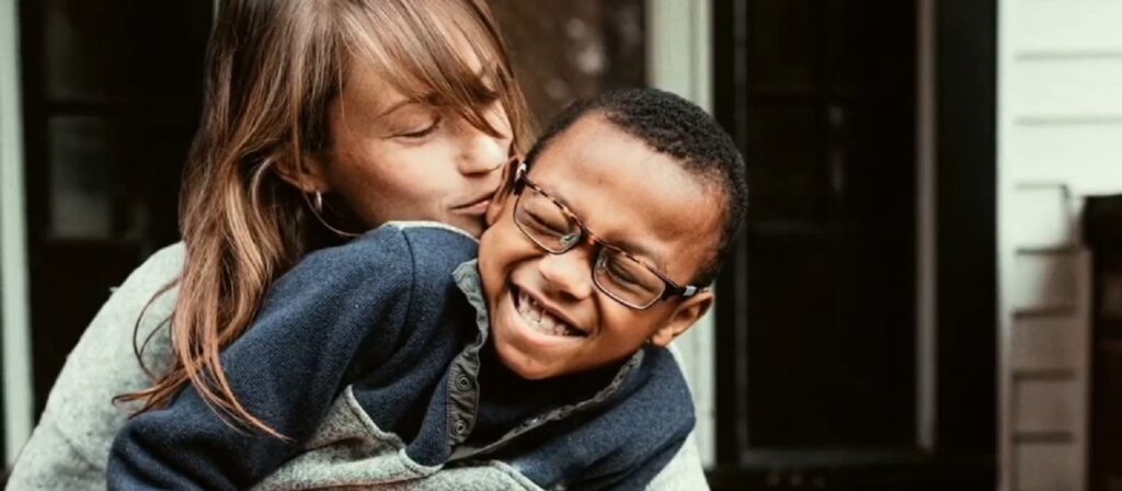 A woman hugs a smiling young boy wearing glasses. They are outdoors in front of a building, and both appear happy and playful.
