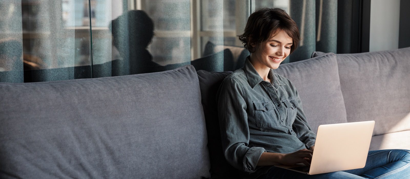 Image of nice young pleased woman using laptop and smiling while sitting on sofa at living room