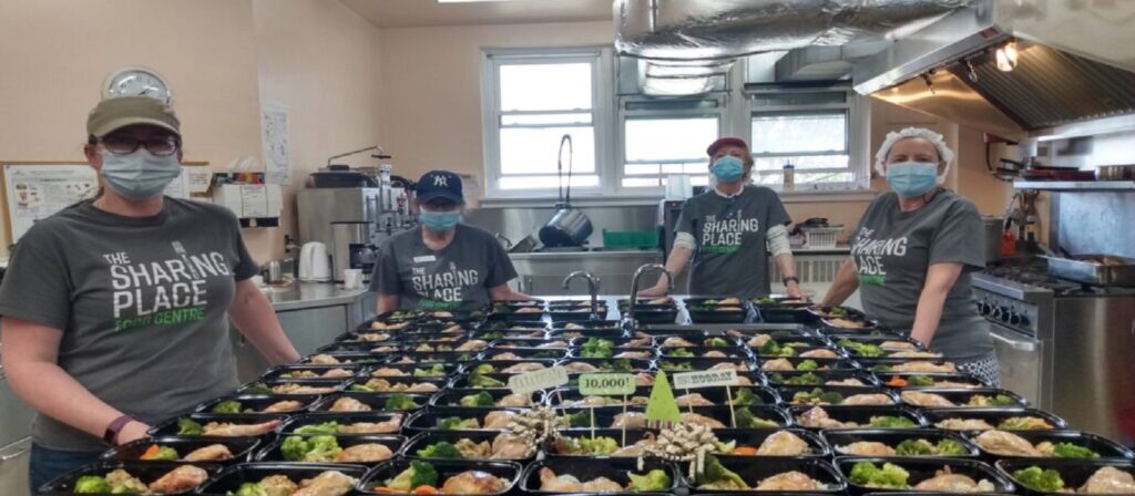 Four volunteers wearing masks and The Sharing Place t-shirts stand in a kitchen behind a counter filled with packaged meals, ready for distribution. Signs with amounts raised are also visible among the meal trays.