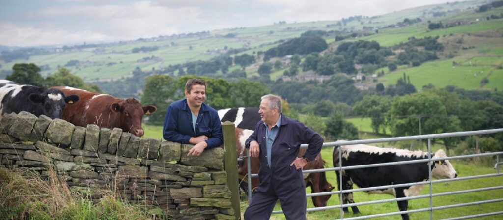 Two men in work clothes stand talking by a stone wall and metal gate in a green countryside field, with several cows grazing nearby and rolling hills in the background.