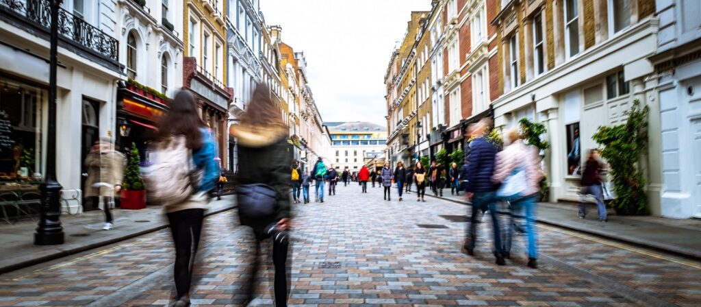 A busy cobblestone street lined with shops and cafes, with people walking in both directions. The buildings are tall, historic, and closely packed. The scene appears lively and vibrant.