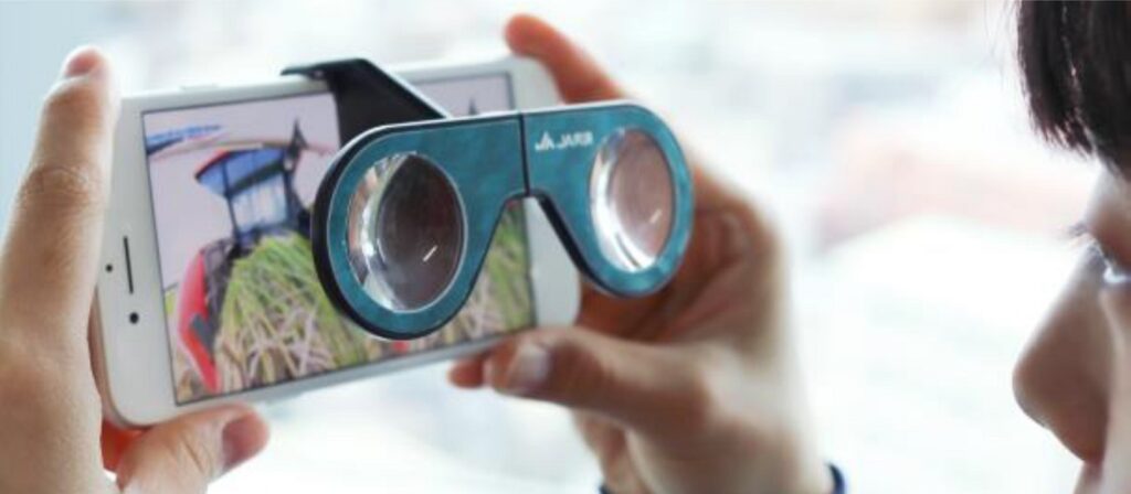A child holds a smartphone displaying an image of a tractor and uses cardboard binoculars to view the screen, creating a virtual reality experience.